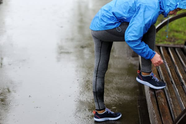 A person wearing a blue rain jacket and grey leggings is seen from the side, bending down to tie their shoe on a wet, wooden bench. The ground is wet, indicating recent rain.