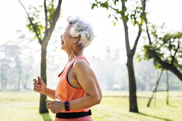 An elderly person with short white hair jogs in a park on a sunny day. They are wearing an orange tank top and a smartwatch. Trees and greenery are visible in the background.