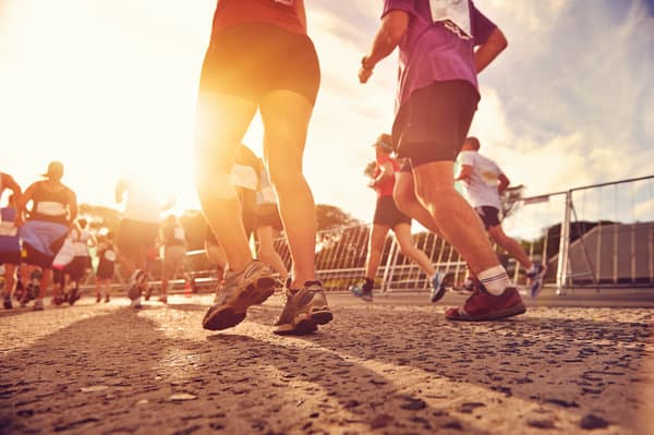 A group of runners is participating in a road race during a sunny day. The image is taken from a low angle, focusing on their legs and running shoes as they move forward on the paved road. The sunlight creates a warm, glowing effect in the background.