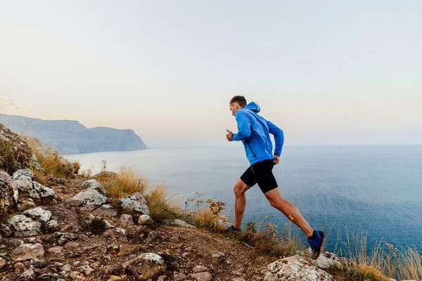 A man wearing a blue jacket and black shorts runs on a rocky trail beside the ocean. The sea and distant cliffs provide a scenic backdrop under a clear sky. The path is lined with patches of grass and scattered rocks.