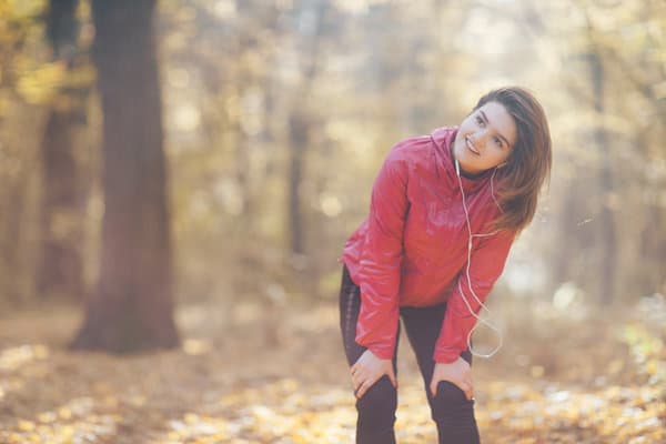 A person wearing a red jacket and earphones is standing outdoors in a forested area, leaning forward slightly with hands on their knees, and looking off into the distance. The background is filled with trees and fallen autumn leaves.