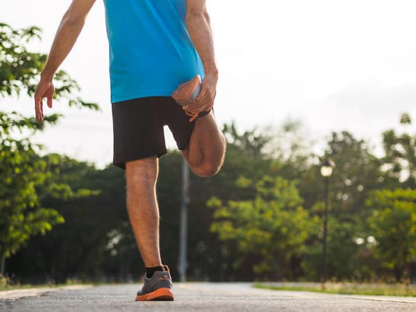 A person in a blue shirt and black shorts stretches their leg by pulling their foot back, standing on one leg on a path in a park. Trees and greenery are visible in the background on a sunny day.