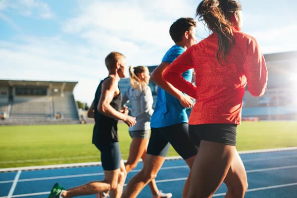 Four athletes are running on a blue track in an outdoor stadium. They are dressed in athletic clothing. The image captures them from behind, with the stadium's empty stands visible in the background under a sunny sky.