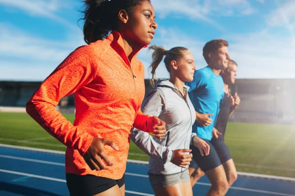 A group of four runners jogs in unison on a blue running track. The foreground features a woman in a bright orange jacket. The background shows a man in a blue jacket, a woman in a light gray jacket, and another man in black athletic gear. The sky is clear and sunny.