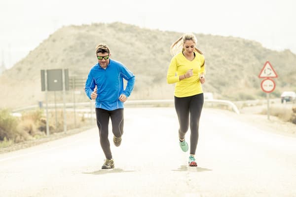 Two people are jogging on an open road with a mountainous backdrop. The person on the left is wearing a blue jacket and black pants, while the person on the right is wearing a yellow jacket and black pants. Both appear to be mid-stride and enjoying the exercise.