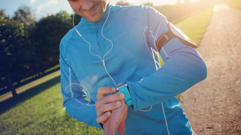 A person wearing a blue athletic outfit and earphones looks at a smartwatch on their wrist. They are outdoors on a sunny day with greenery in the background and sunlight casting a warm glow. The person appears to be in a park or on a running path.