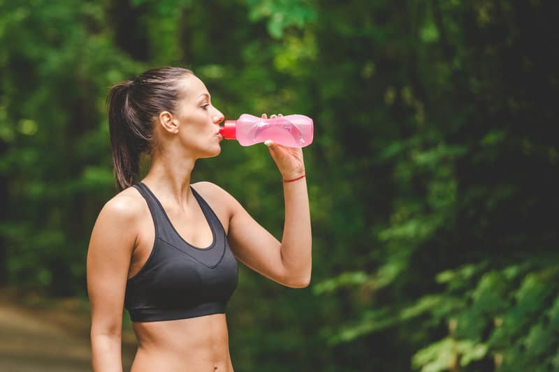 A person with a ponytail, wearing a black sports bra, is drinking from a pink water bottle while standing outdoors in a wooded area. Trees and greenery surround the scene.