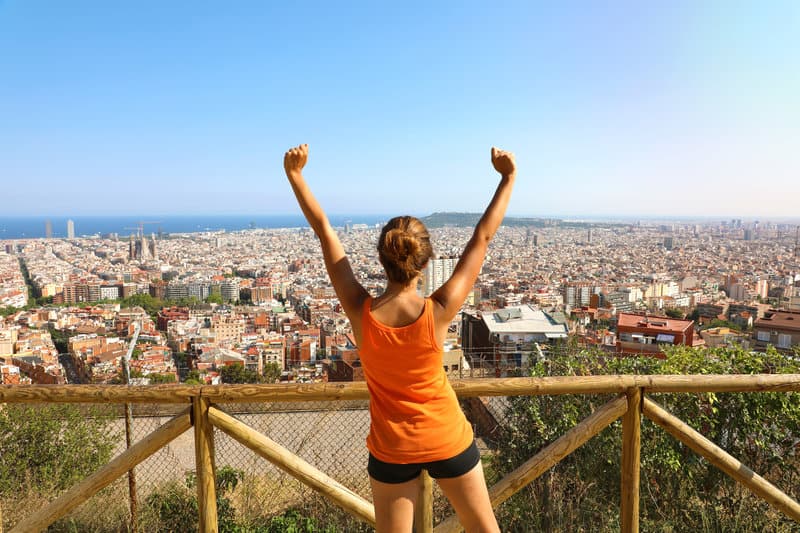 A woman stands with her back to the camera, raising her arms in triumph, overlooking a panoramic view of a sprawling city with buildings, mountains, and the sea in the distance. She is wearing an orange tank top and black shorts, standing by a wooden fence.