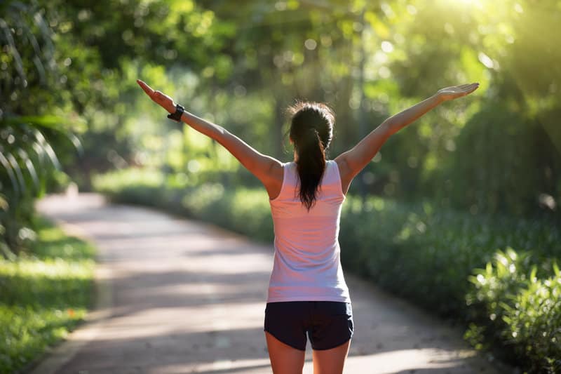 A woman in athletic wear stretches her arms wide while walking on a sunlit path in a lush, green park. She is wearing a white tank top and dark shorts, with her back to the camera. The scene is bright and serene, with sunlight filtering through the trees.