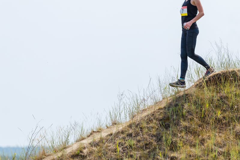 A person in athletic clothing runs along the top of a grassy hill, with a race bib visible on their chest. The background is a clear, pale sky, and the terrain is sparsely vegetated with grass. The scene suggests participation in an outdoor running event.