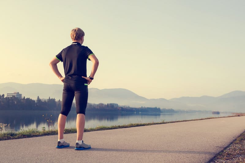 A person stands on a paved path with hands on hips, facing a scenic view of a river surrounded by mountains under a clear sky. They are wearing athletic clothing, including shorts, a shirt, and running shoes.