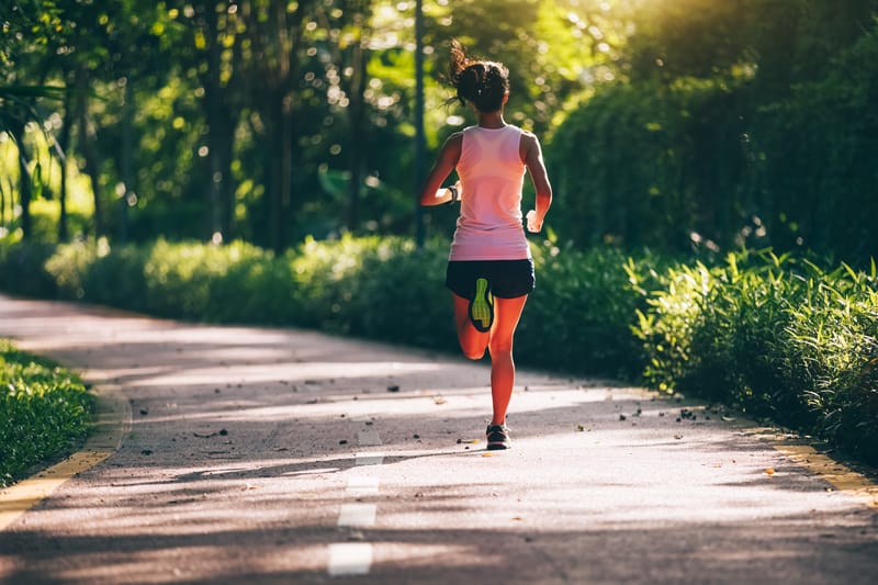 A person wearing a white tank top and dark shorts is jogging along a paved path through a lush, green park. The path is lined with trees and sunlight filters through the foliage, creating a serene and refreshing atmosphere.