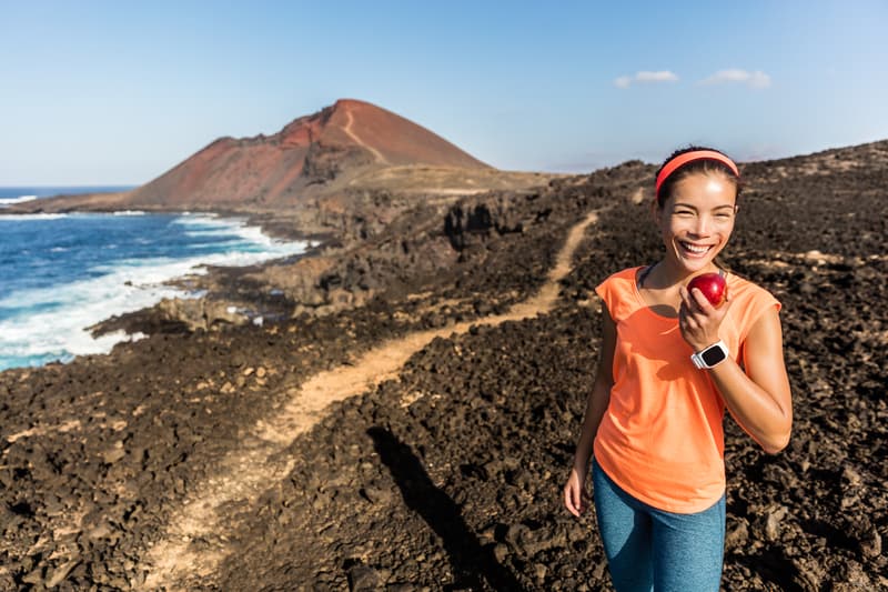 A smiling woman in activewear stands on a rocky coastal path, holding an apple. She wears an orange top, blue leggings, and a smartwatch. In the background, there is a rugged landscape with a red hill and ocean waves crashing against the rocks under a clear blue sky.