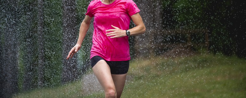 A person wearing a bright pink shirt and black shorts is running outside in the rain. The water droplets are visible in the air, creating a dynamic scene of motion. The background is blurred with green foliage.