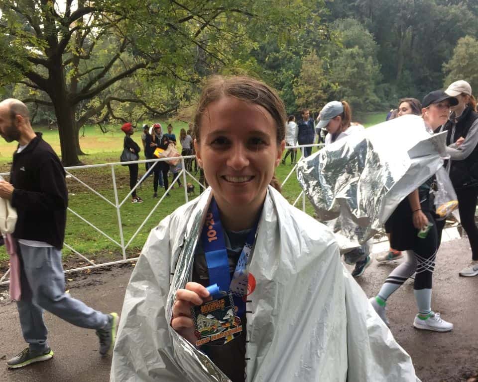 A person wrapped in a silver foil blanket smiles while holding up a marathon medal. They are outdoors in a park, surrounded by other people and trees. The atmosphere suggests a recently concluded race event.