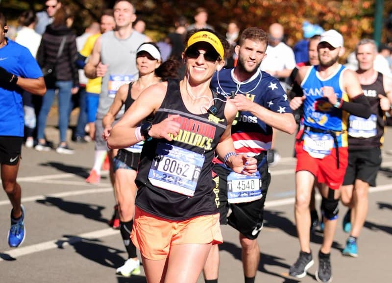 A group of runners in athletic gear participates in a road race. The runner in the foreground, wearing a black tank top, orange shorts, and a yellow visor, appears to be smiling and giving a thumbs-up. Other runners are visible in the background, focused on the race.