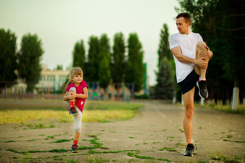 A man and a young girl are exercising outdoors on a track. The man is dressed in a white t-shirt and black shorts, while the girl wears a pink top and white leggings. Both are lifting one leg in a stretch. Trees and buildings are visible in the background.