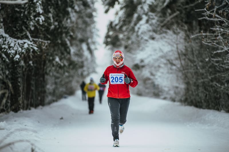 A runner wearing a red jacket, gray pants, and a numbered bib 205 is jogging along a snow-covered trail surrounded by snow-laden trees. Other people can be seen in the background. The scene is wintery and serene.