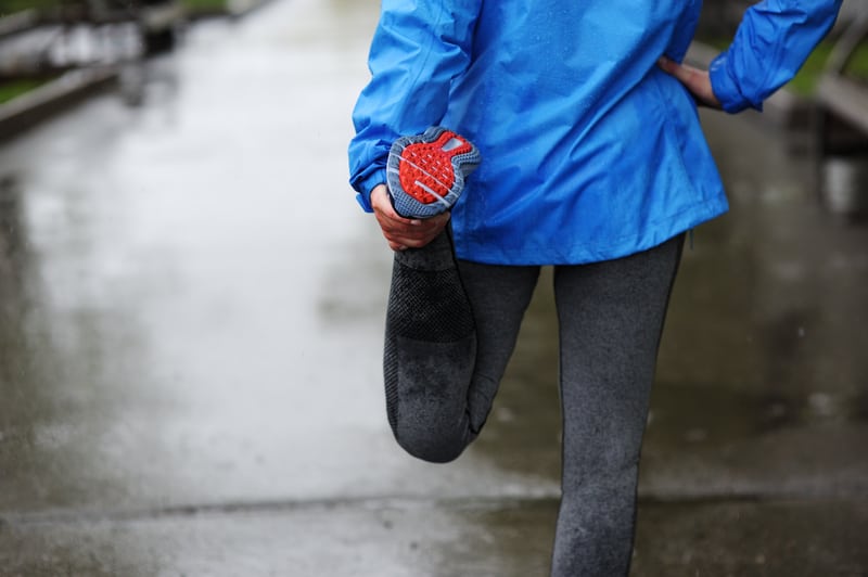 A person wearing a blue rain jacket and gray leggings stretches their leg by holding their ankle behind them on a wet pavement. The scene appears to be outdoors on a rainy day, likely preparing for a run. Only the lower half of the person's body is visible.