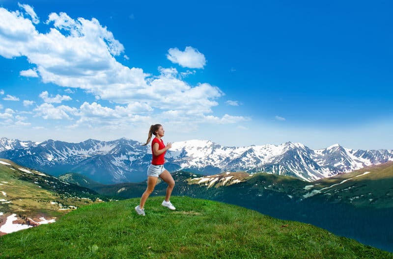 A woman wearing a red shirt and grey shorts is running on a grassy hilltop with a backdrop of snow-capped mountains under a bright blue sky with scattered clouds.