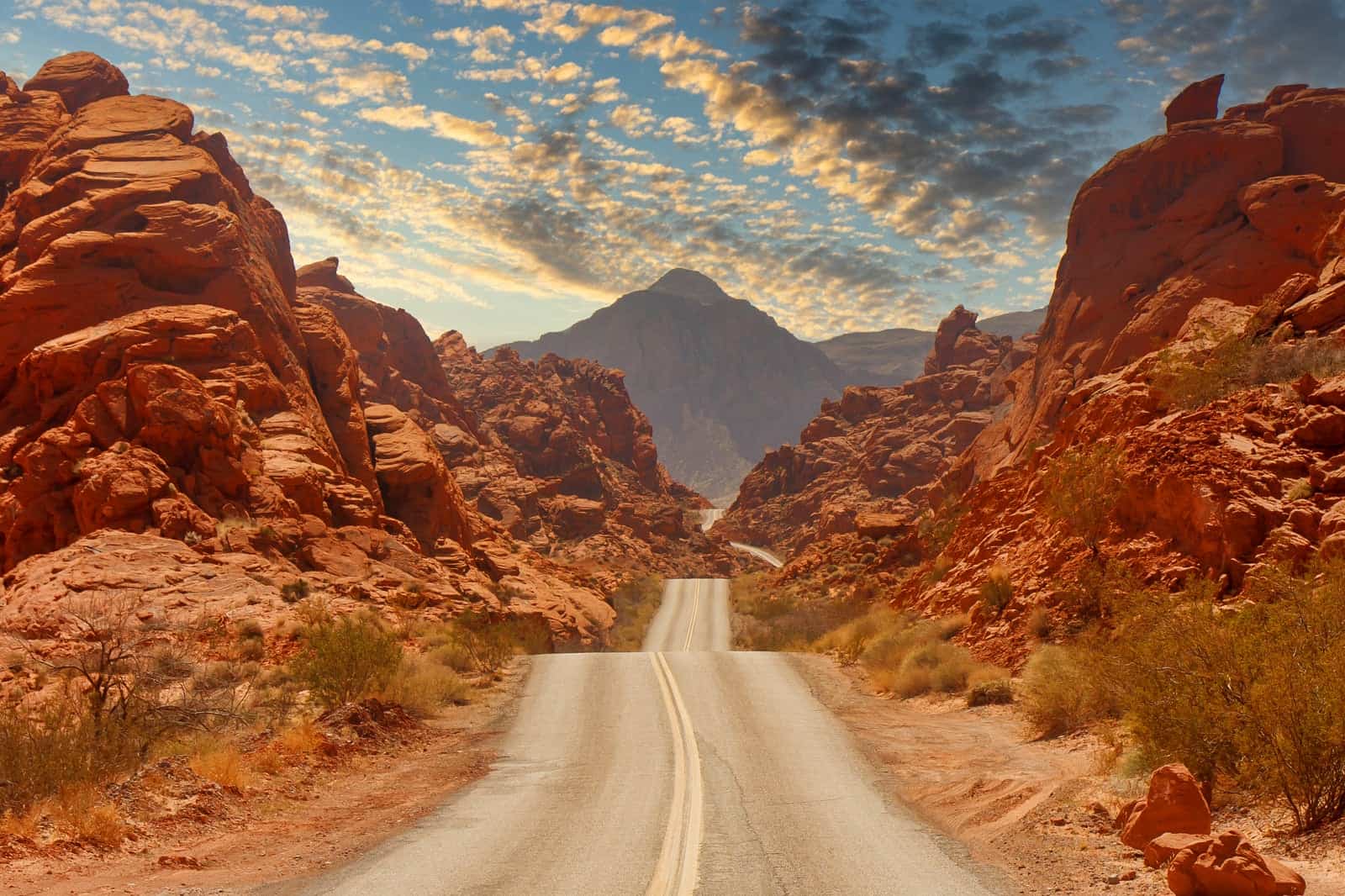 A winding road passes through red rocky formations under a partly cloudy sky with a distant mountain peak. The rugged terrain and dramatic landscape create a striking desert scene, with sparse vegetation lining the road—a perfect backdrop for those who travel to compete in half marathons.