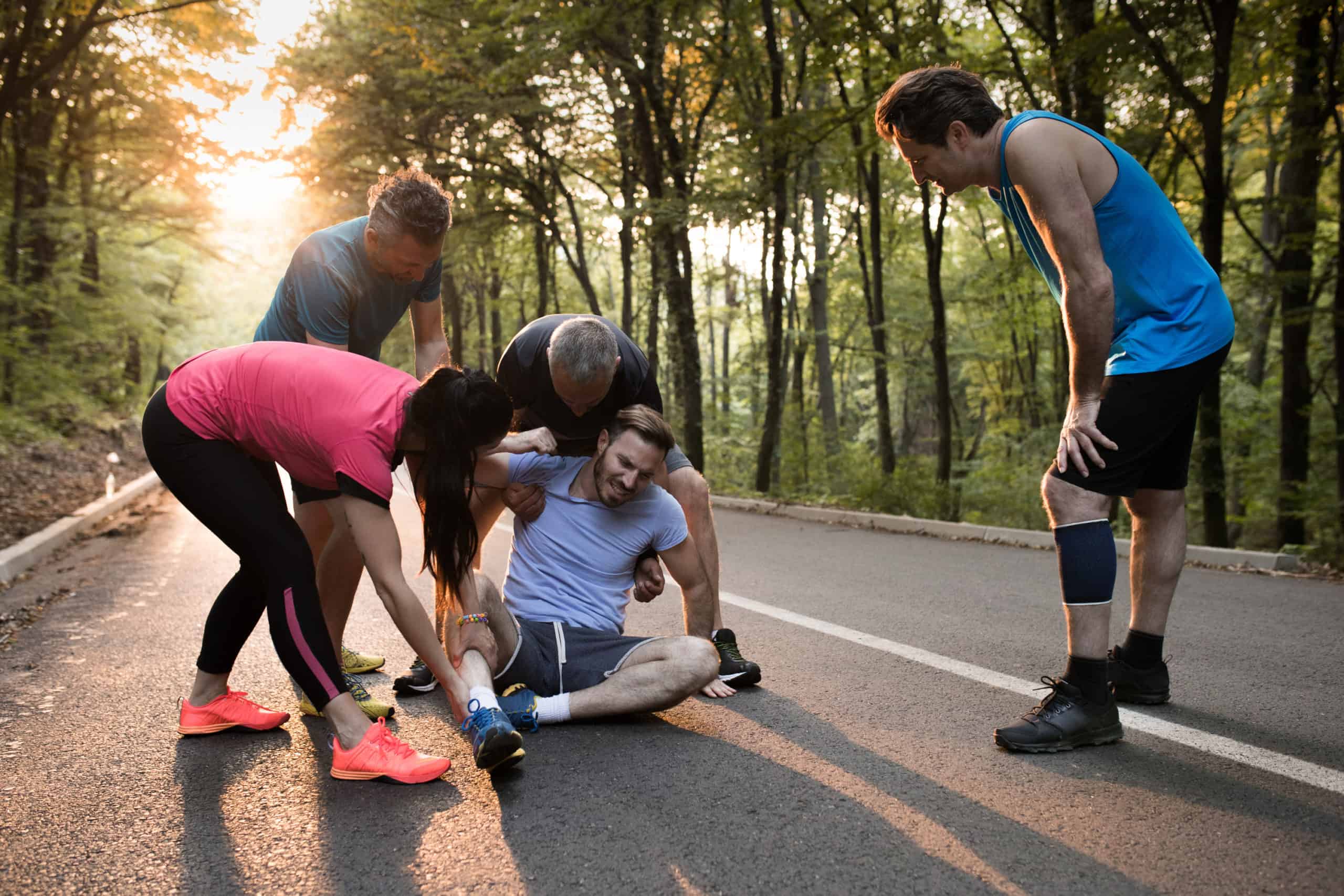 A group of five adults in athletic wear are on a forest road. Two people are helping a seated man who appears to be injured, while two others stand nearby, observing and showing concern. The sun is setting or rising in the background, casting a warm glow through the trees.