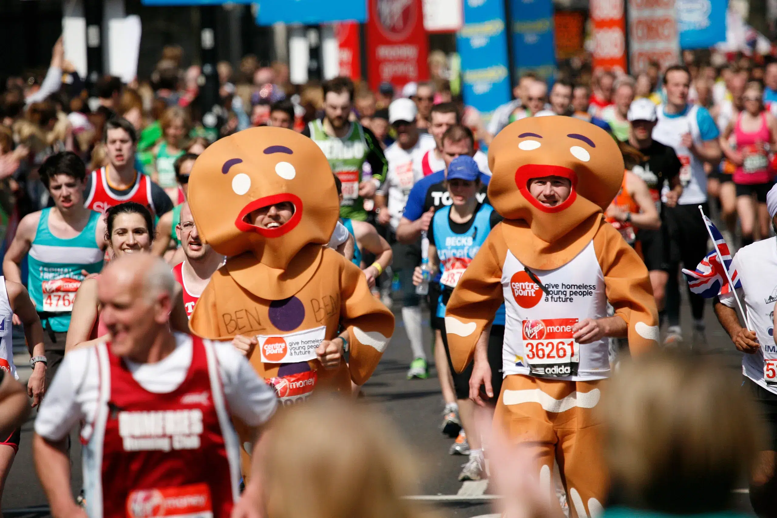 Two participants dressed as gingerbread men run among a crowd of runners in a Halloween half marathon. One costume has "BEN R" written on it. Spectators line the route, and various colorful banners and signs are visible in the background.