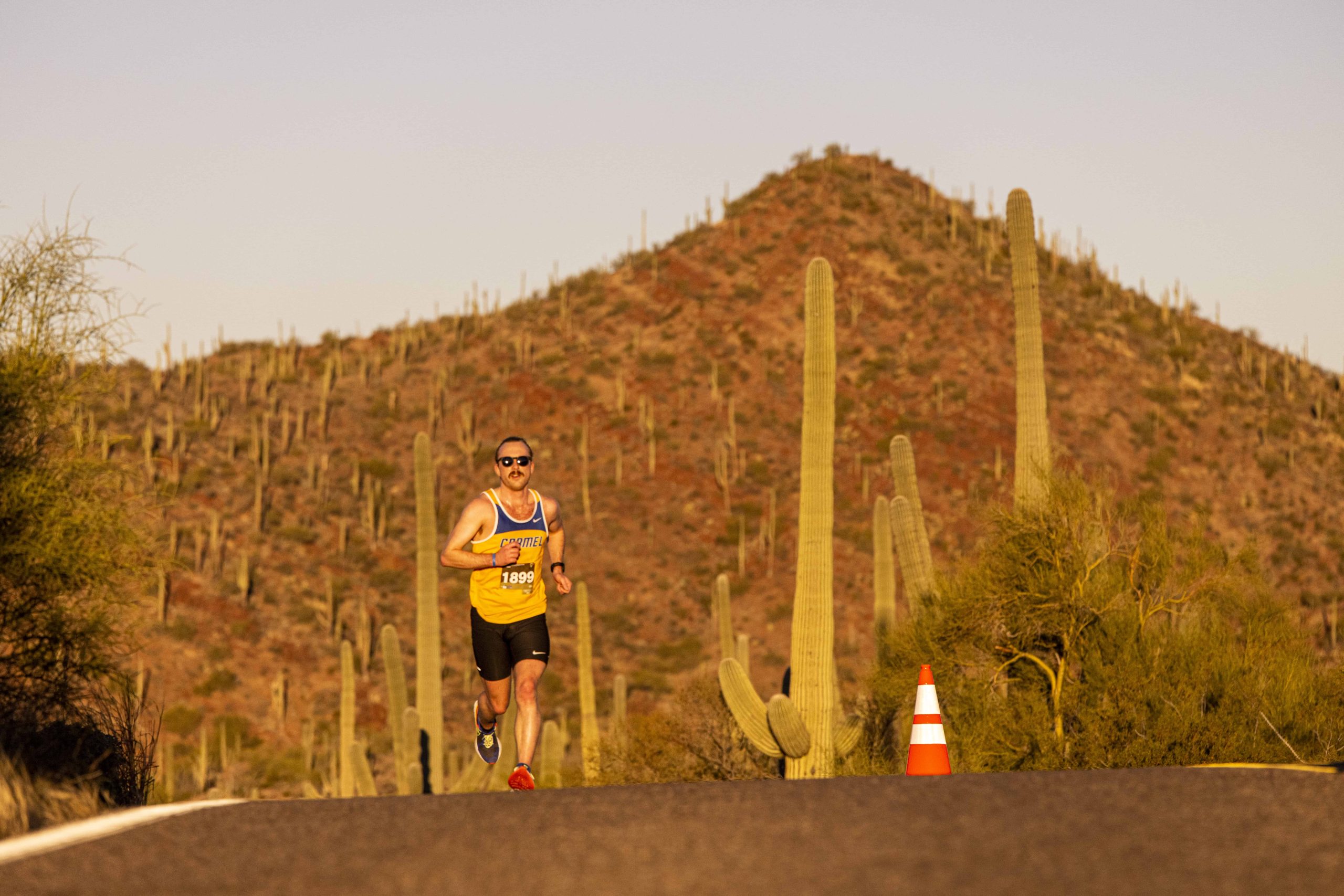 A person runs on a paved road in a desert landscape with tall cacti and a distant hill under a clear sky. The runner, participating in the Saguaro Half Marathon, wears a yellow tank top, black shorts, and sunglasses, and is numbered 1290. An orange traffic cone is seen on the side of the road.