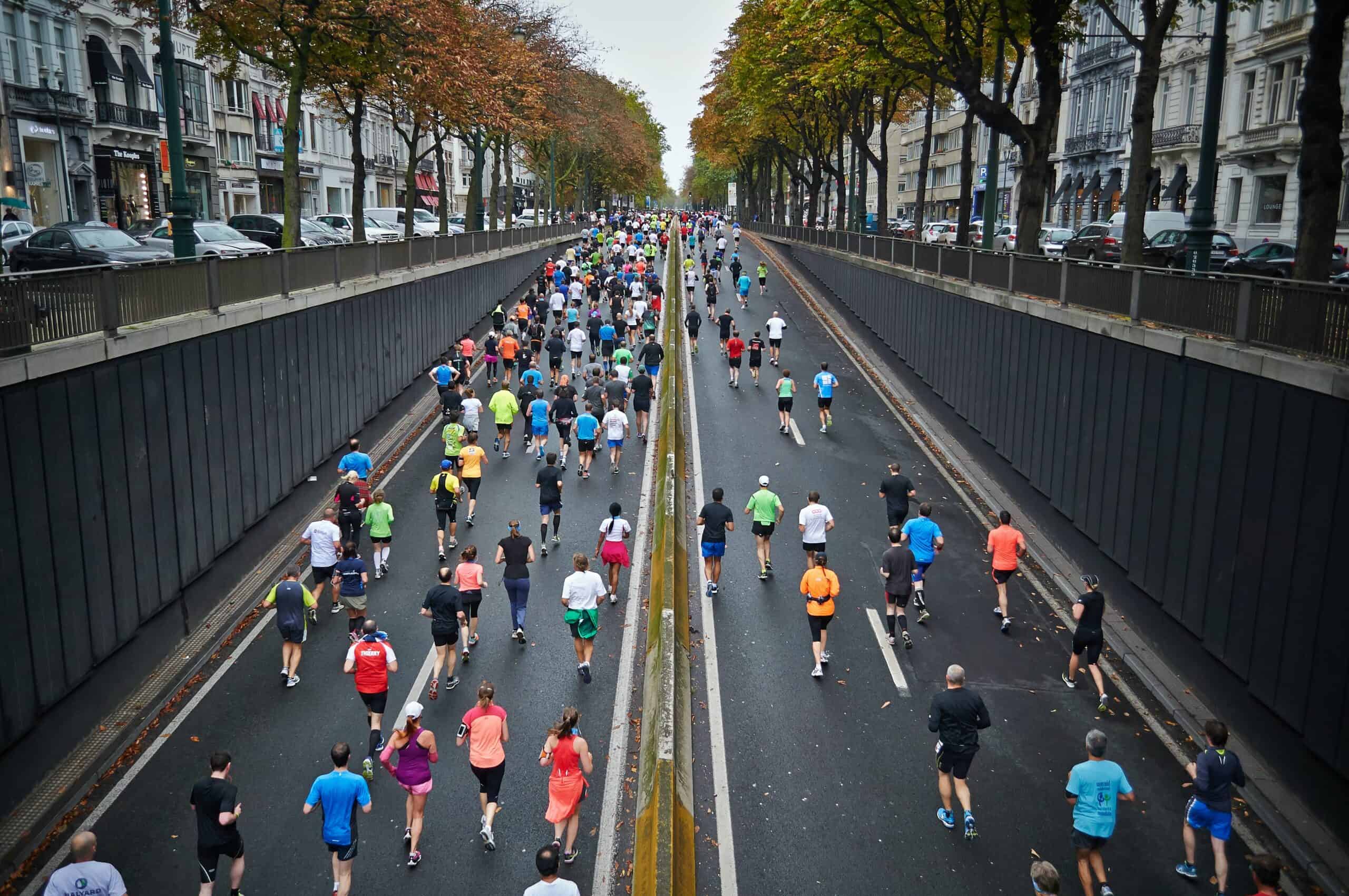 A large group of people in colorful sportswear are running a half marathon on a city street with lanes separated by a median. Tall buildings and trees line both sides of the road, and some spectators can be seen in the distance under cloudy skies.