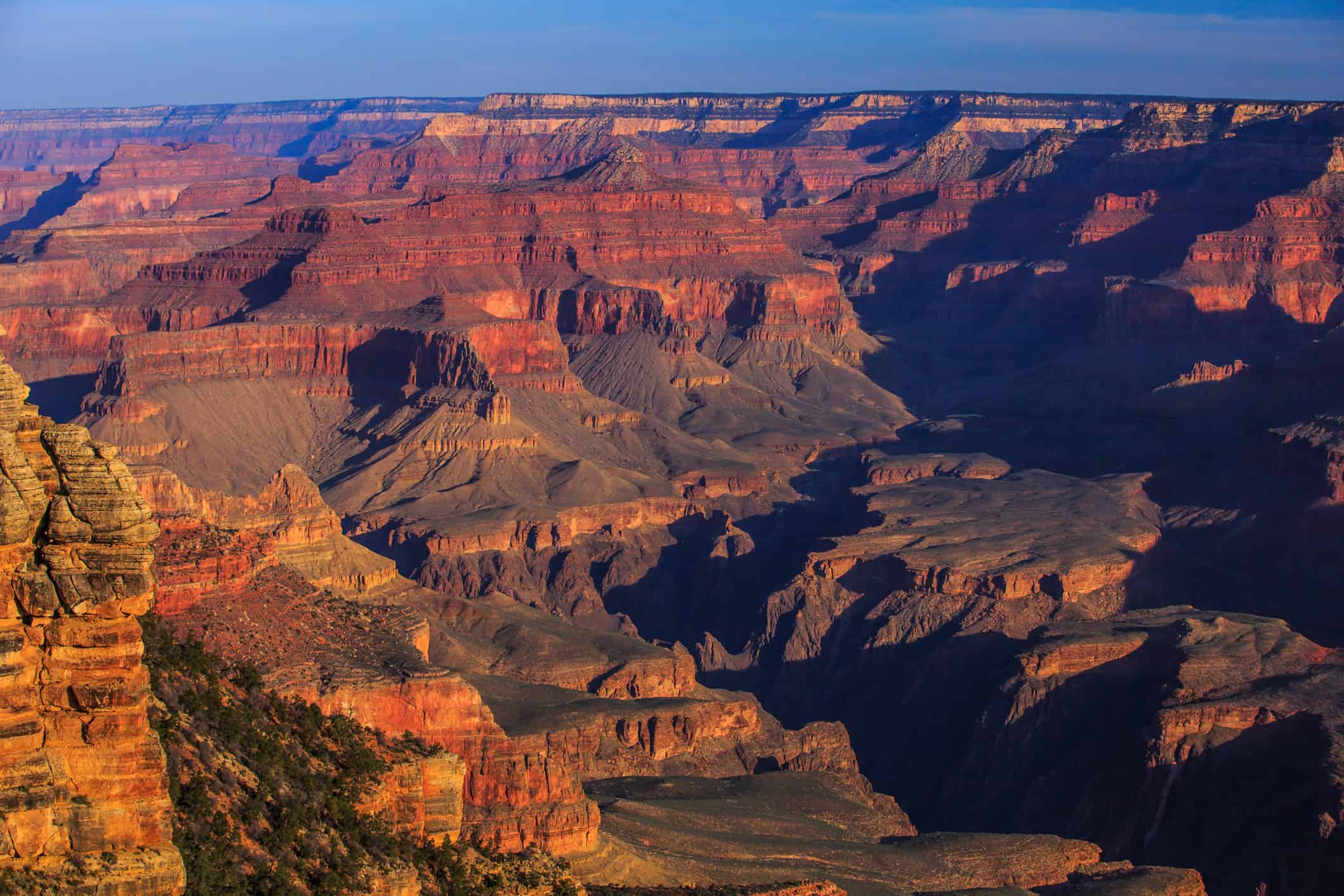 A sweeping view of the Grand Canyon, showcasing its rugged rock formations and deep, expansive valleys. Layers of red, orange, and brown hues highlight the geological strata. The scene is bathed in warm sunlight with clear blue skies overhead.