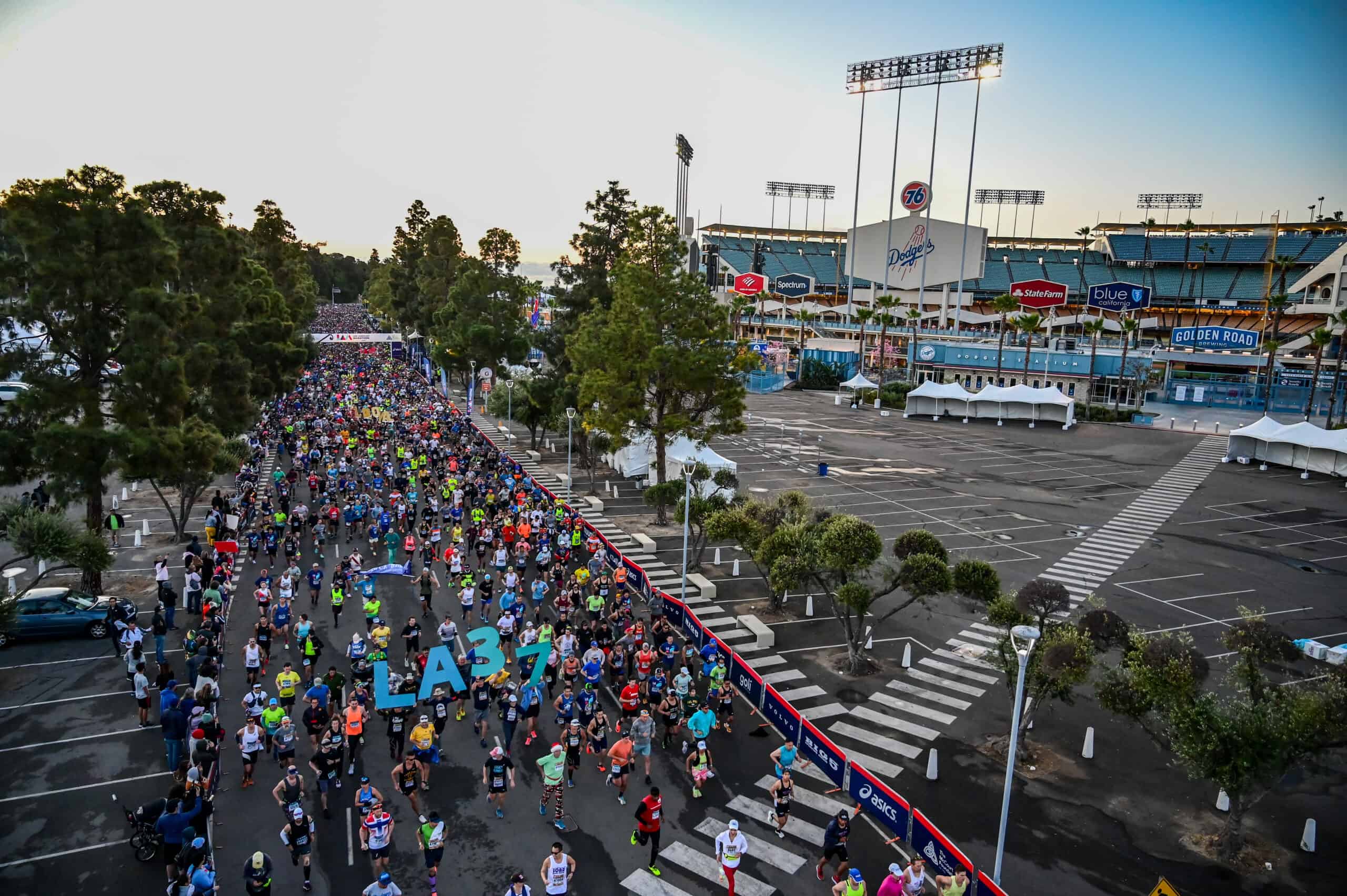 A large crowd of marathon runners starting a race near a stadium with "LA37" marking the event. The stadium, visible in the background, features logos of various sponsors. The street is lined with trees, and early morning light illuminates the scene.