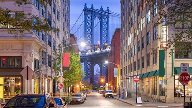 A nighttime street view in New York City's Dumbo neighborhood, featuring the Manhattan Bridge framed by buildings on either side. The scene includes parked cars, lit streetlights, and a stop sign in the foreground. The bridge's structure is illuminated, standing out against the twilight sky, reminiscent of Brooklyn half marathons' scenic routes.