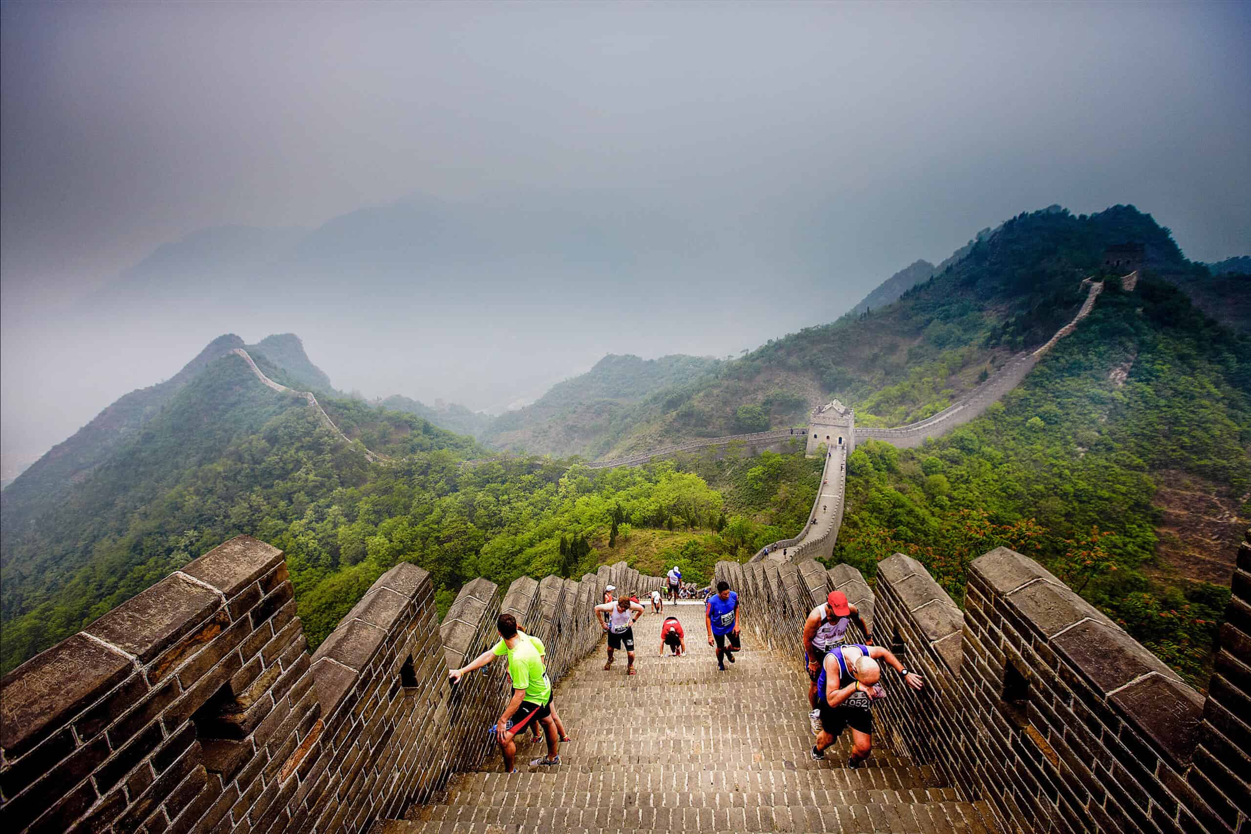 Tourists climb a steep section of the Great Wall of China, surrounded by mist and lush green hills. The ancient structure stretches into the distance, winding through the rugged landscape under a cloudy sky, reminiscent of adventurous half marathons that challenge and inspire.