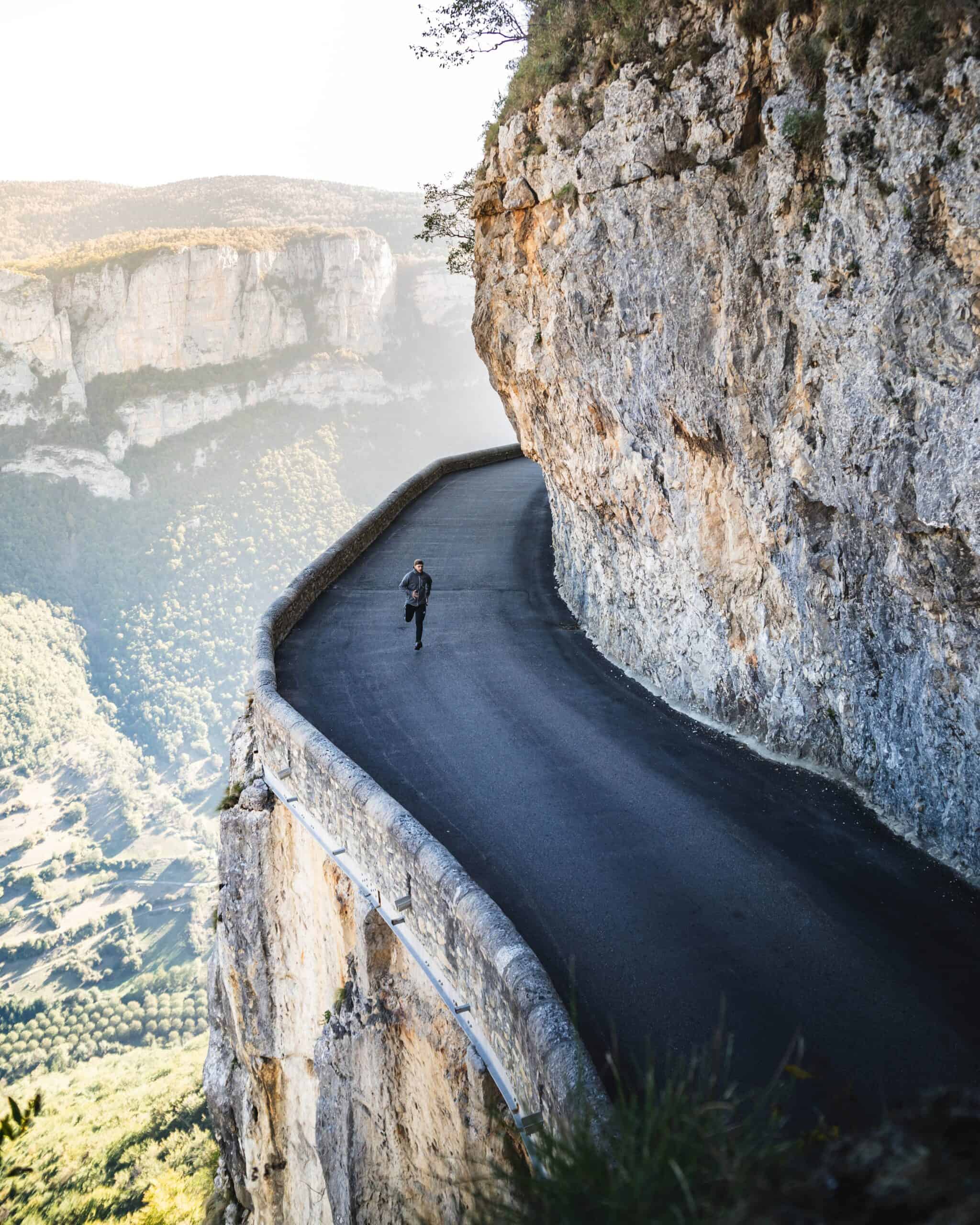 A person jogs along a narrow, winding mountain road cut into the side of a steep cliff, with a picturesque landscape of green valleys and rocky mountains in the background. Focused on their running goals, the runner continues past the stone barrier that borders the cliff side.