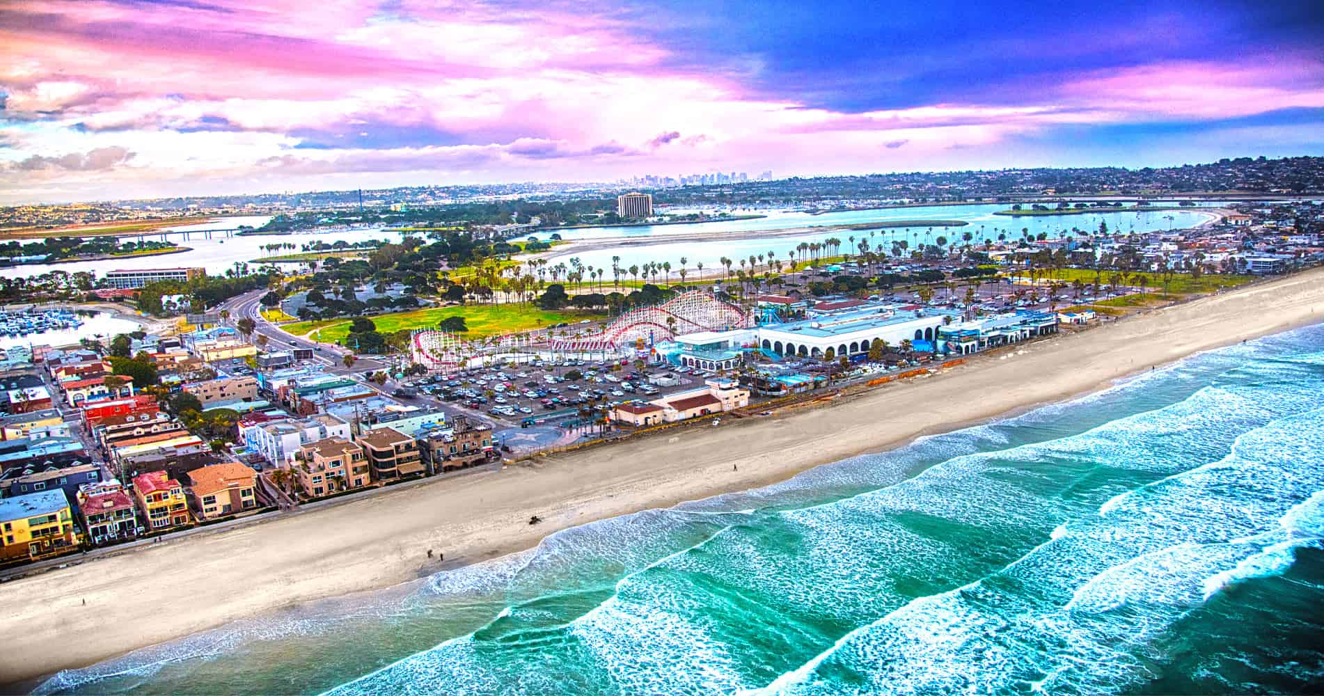 A scenic aerial view of a coastal city at sunset captures the essence of San Diego. The image shows a long beachfront with waves crashing onto the shore, colorful buildings and a rollercoaster near the beach, and a series of water bodies and green areas further inland under a vibrant sky, perfect for San Diego half marathons.