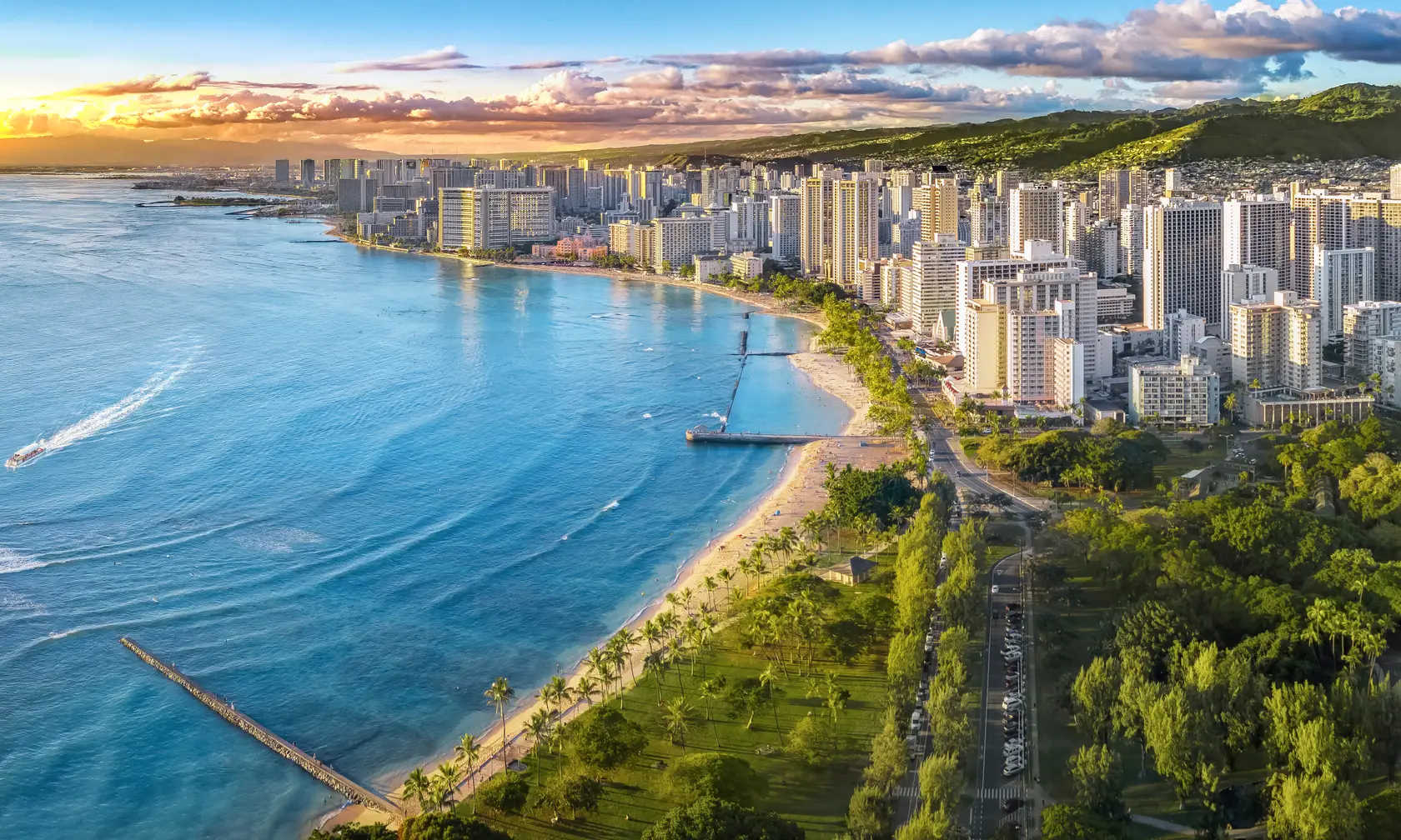 Aerial view of Waikiki Beach in Honolulu, Hawaii at sunset. High-rise buildings line the coast, with lush green parks and palm trees along the beachfront. The calm blue ocean stretches out to the horizon, and clouds dot the sky.