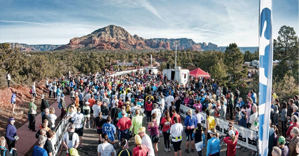 A large, colorful crowd of runners gathers at the start line of an outdoor race event. The scene is set in a scenic, mountainous area with red rock formations and trees. Known as one of the most scenic half marathons, the sky is clear, and there are tents and event signage in the background.