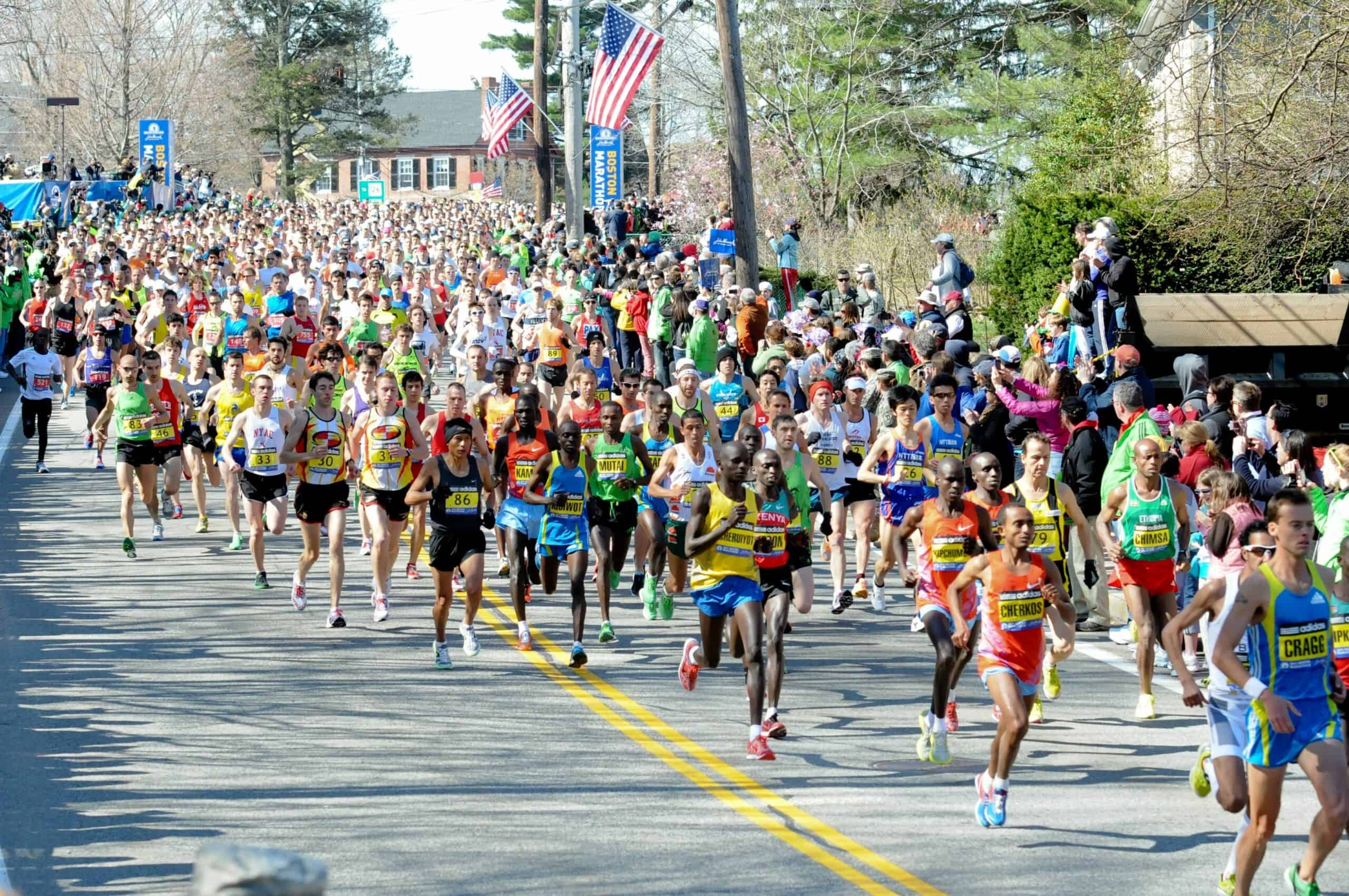 A large, diverse group of runners participating in a marathon on a road lined with spectators. The runners wear colorful athletic gear. American flags are visible in the background, and spectators stand along the sides cheering. Trees and houses line the route.