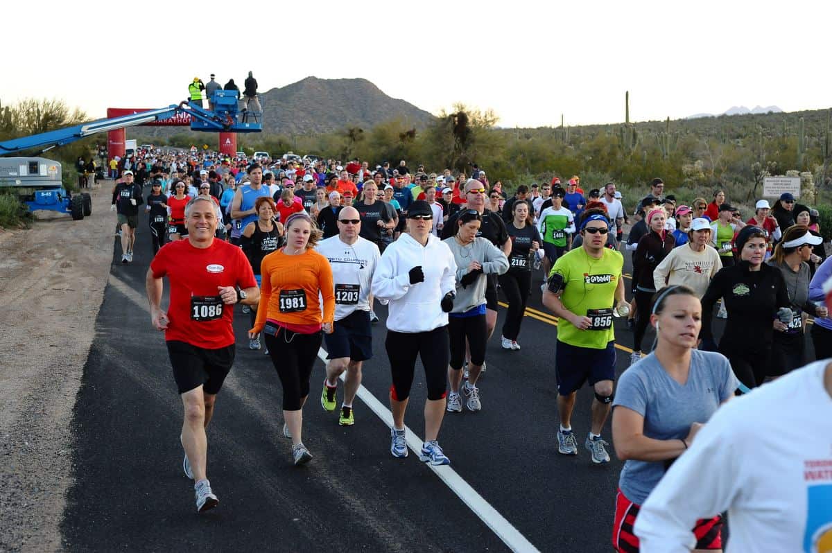 A large group of runners participating in one of the Phoenix half marathons on a paved road through a desert landscape. They are wearing race bibs and various athletic gear. In the background, there are mountains, cacti, and some spectators near the starting area.