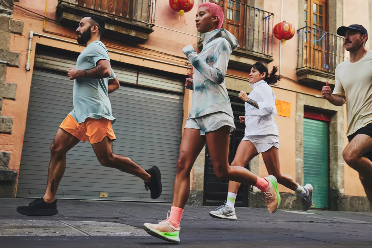 A group of four people jogs down a street in an urban setting with colorful, closed storefronts in the background. The weather appears mild, and they all wear athletic attire from lululemon running gear, including pastel-colored shorts, hoodies, and running shoes.