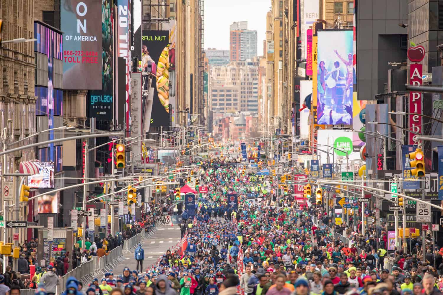 A large group of marathon runners fills a street in an urban setting with tall buildings and billboards lining both sides. Known for hosting popular half marathons, the crowd appears diverse and colorful, with various signs and traffic lights visible above. The atmosphere is busy and energetic.