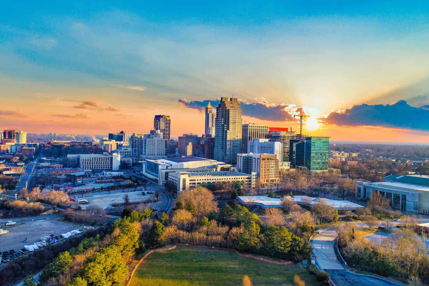 Aerial view of a city skyline at sunset with a mix of tall buildings and shorter structures. The sun is partially hidden behind clouds, casting a warm glow. Green spaces are visible in the foreground, dotted with trees and open areas, perfect for training routes like Raleigh half marathons.