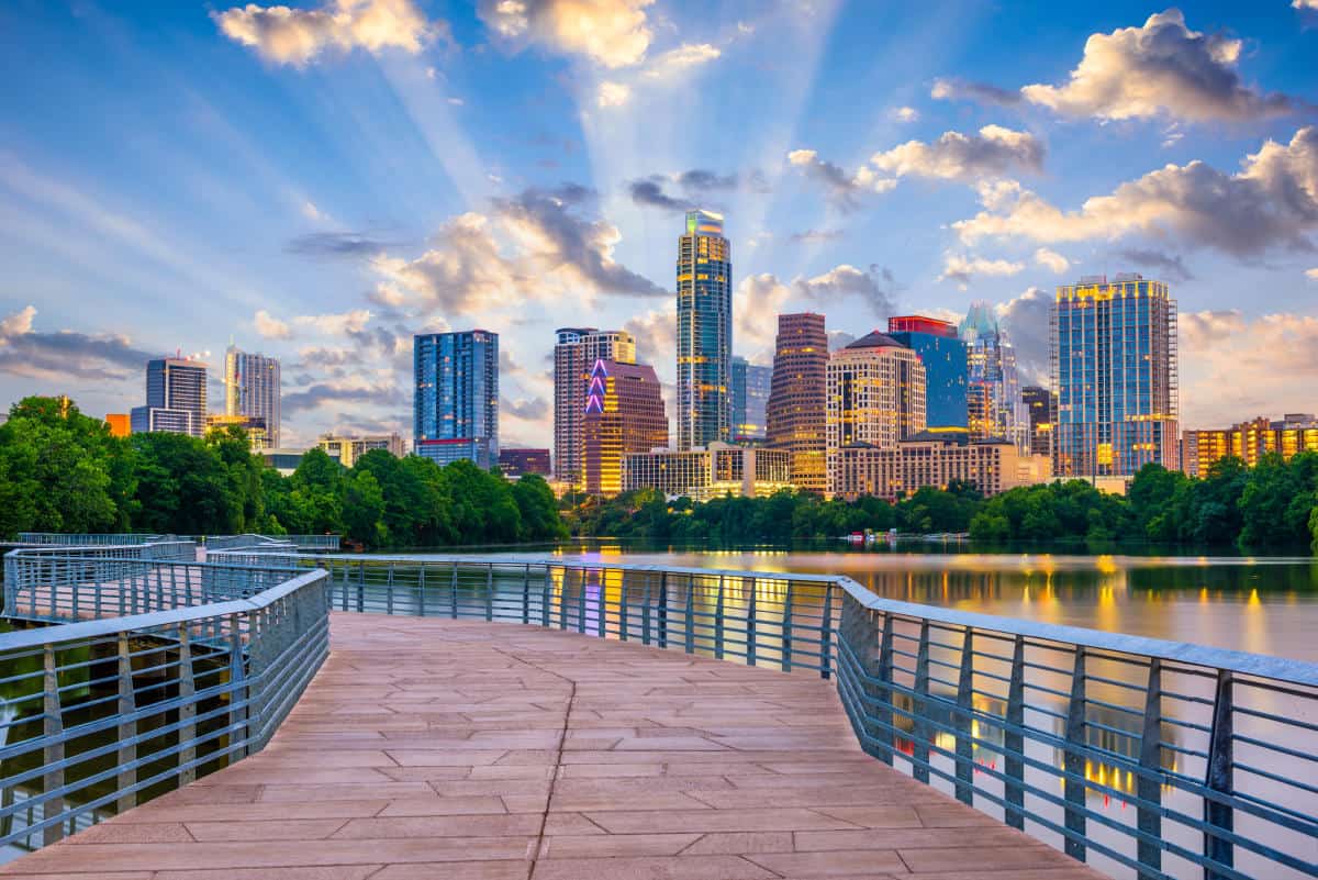 A vibrant city skyline with tall buildings is seen across a calm body of water in Austin. The sky is partially cloudy with sun rays peeking through. A wide boardwalk with metal railings leads toward the water, surrounded by lush green trees on either side, perfect for a scenic half marathon route.