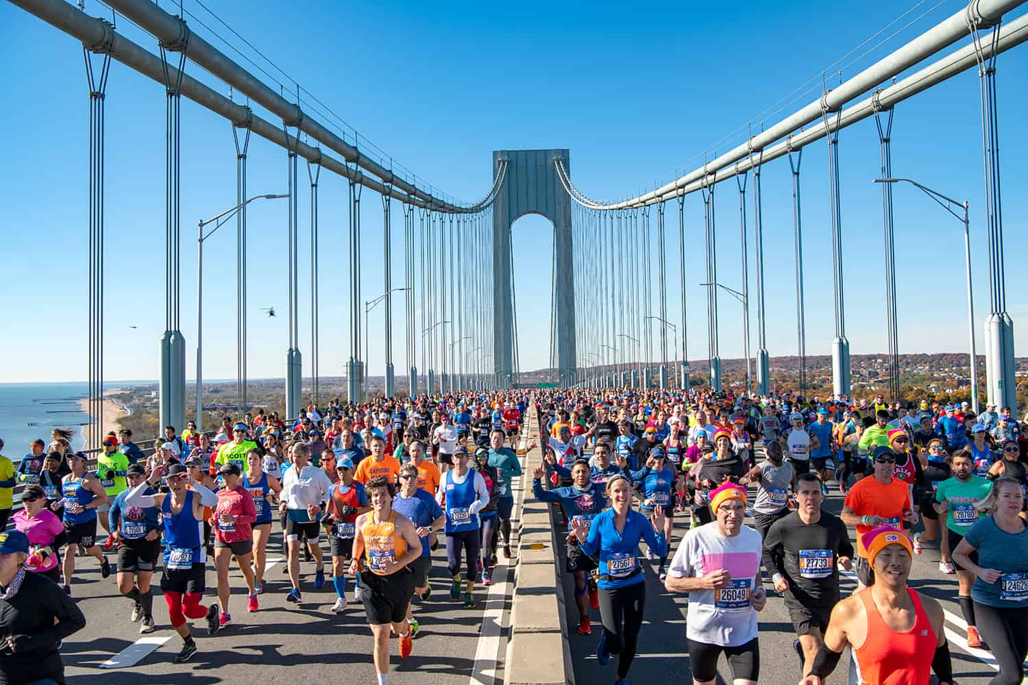 A large group of marathon runners, part of the Abbott World Marathon Majors, is crossing a wide suspension bridge. The sky is clear and blue, and the sea is visible to the left. The bridge towers can be seen in the background, and the participants are in colorful athletic gear.