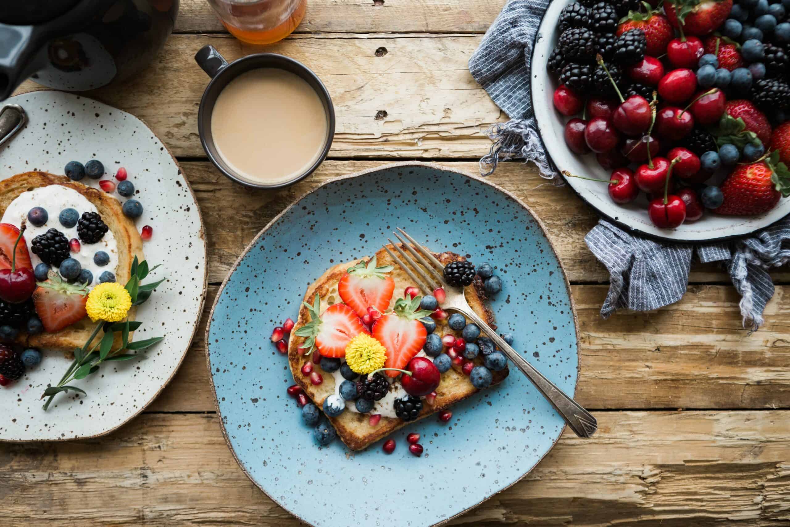 A rustic wooden table hosts a runner's breakfast with a blue plate holding toast topped with yogurt and assorted fresh berries, a fork beside it. A cup of coffee, a bowl filled with various berries, and another plate with similar toast are arranged around the main plate.