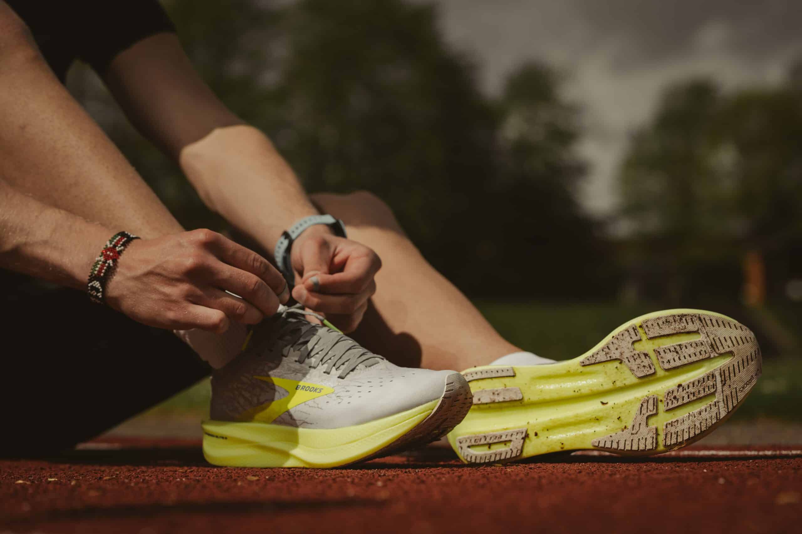 A person sitting on a running track lacing up light gray and neon yellow running shoes, possibly after a half marathon recovery. The image focuses on the shoes and hands, with a blurred background of green trees under a cloudy sky. The person wears bracelets and a watch.