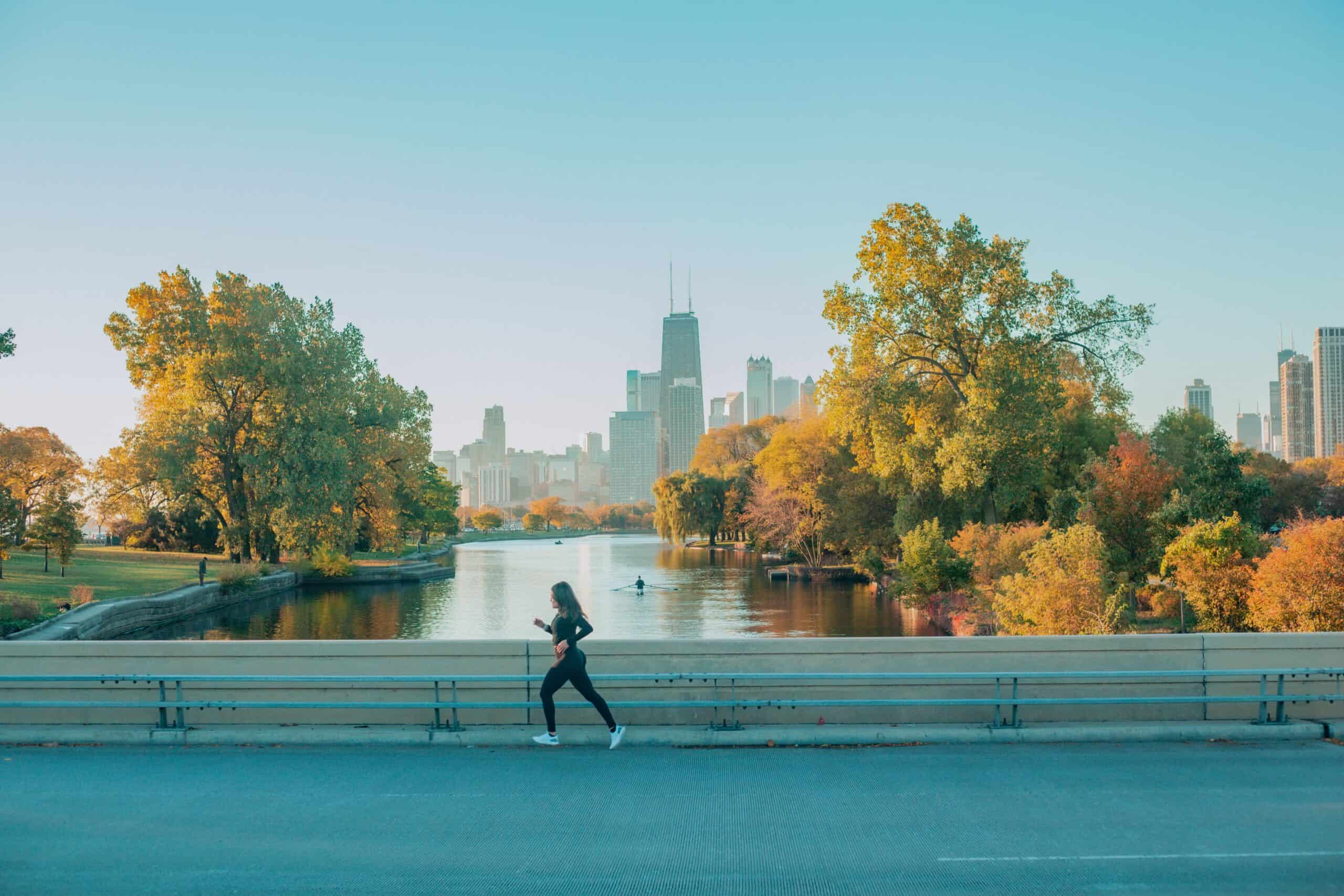 A person jogs along a barrier-lined road with a river, trees, and the Chicago skyline, including a tall building, in the background. The scene is bathed in soft sunlight, giving it a warm and vibrant feel, making their run truly picturesque.