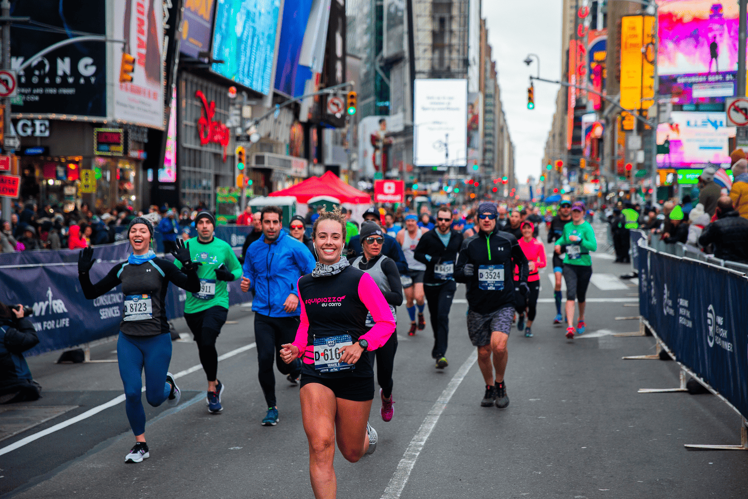 Runners participate in a marathon through a bustling city street lined with tall buildings and bright billboards. The lead runner, wearing a pink and black outfit, is smiling. Spectators stand behind barriers on the side of the road, cheering on the participants.