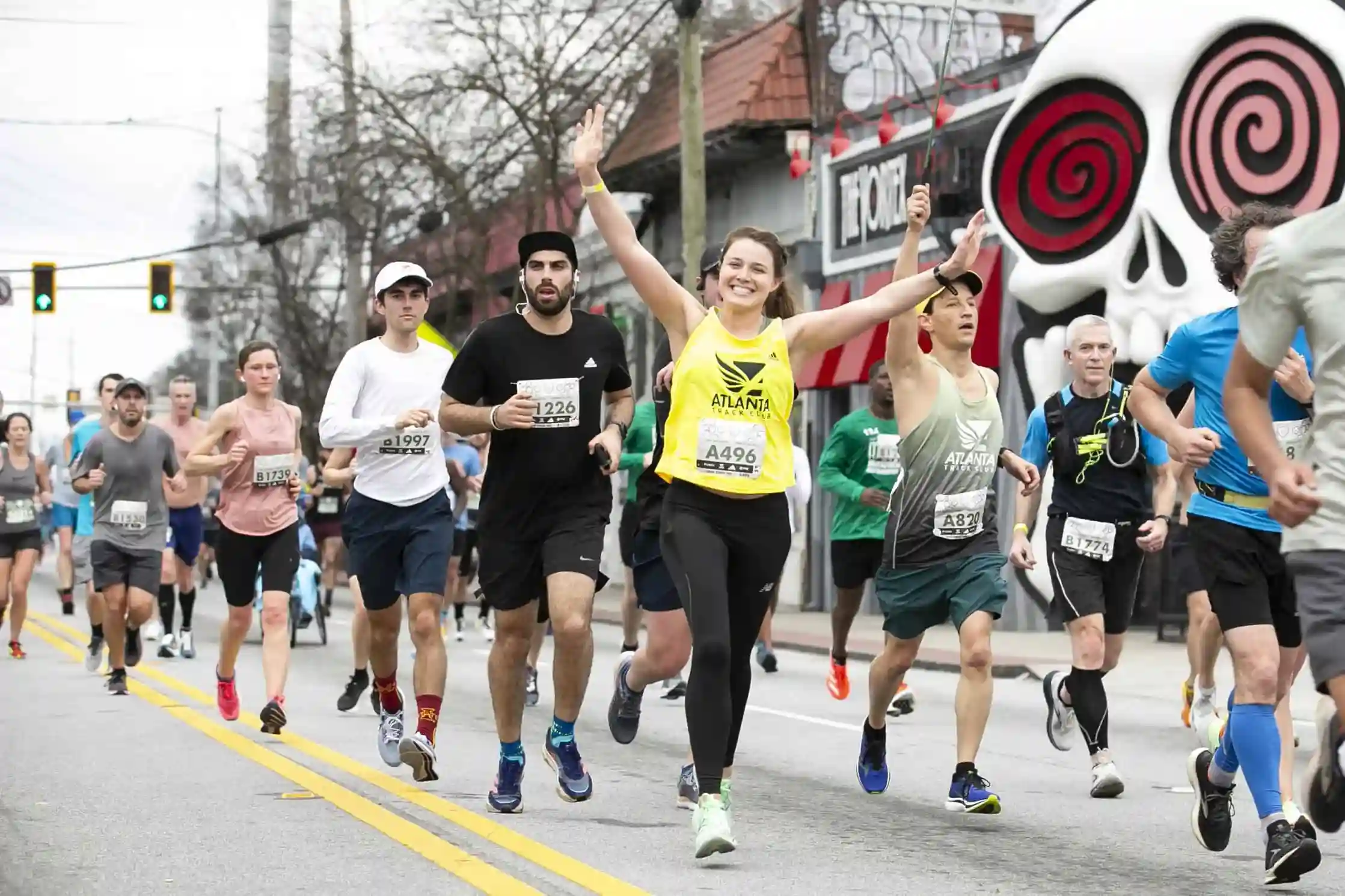 A group of runners participating in a race on a city street. A woman in a yellow tank top raises her arms cheerfully. Graffiti art featuring a large skull with red eyes is visible on a building in the background. Other runners, traffic lights, and trees are present.